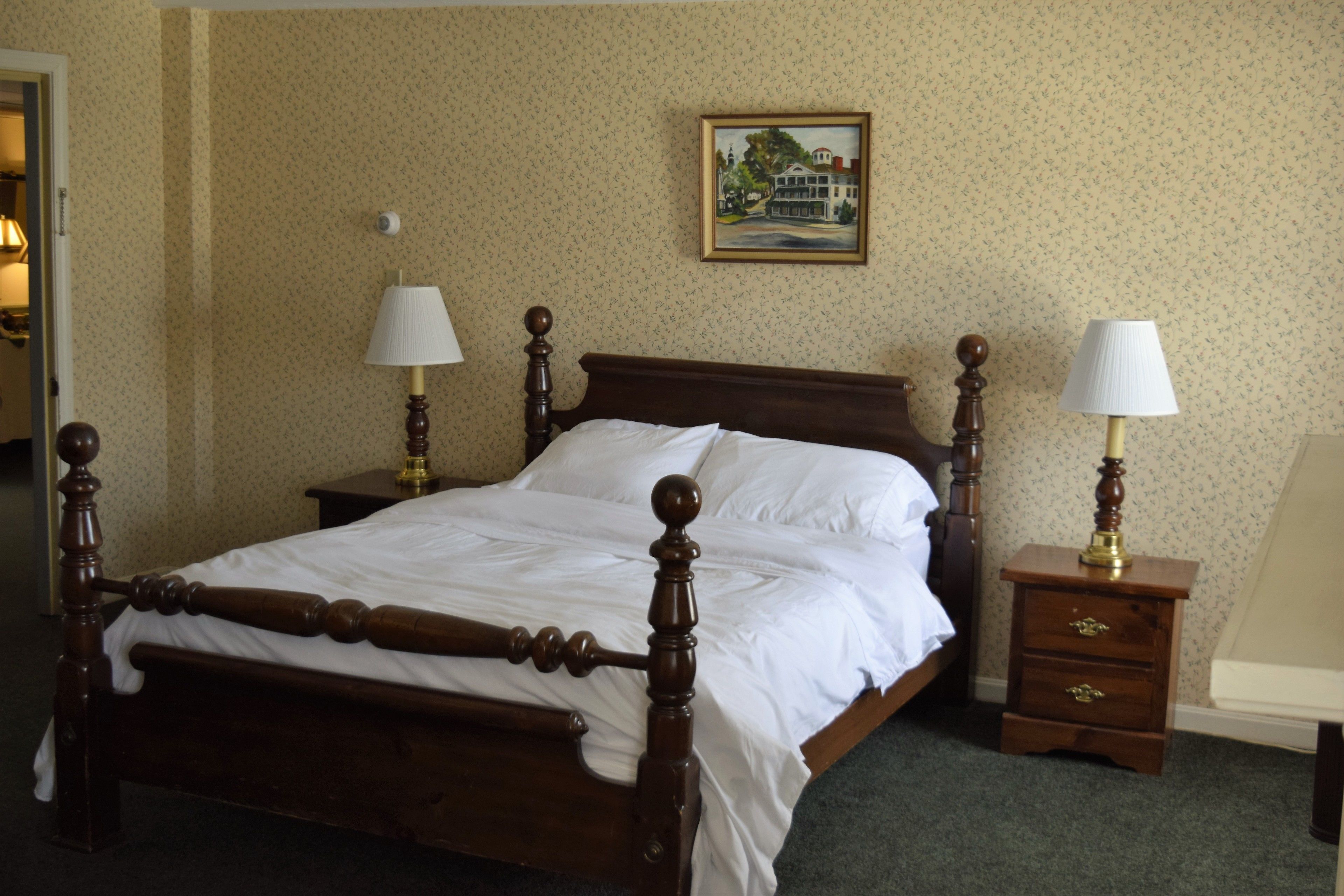 A neatly made bed with a wooden frame and white linens, two bedside tables with lamps, a framed picture on the wall, and a carpeted floor.