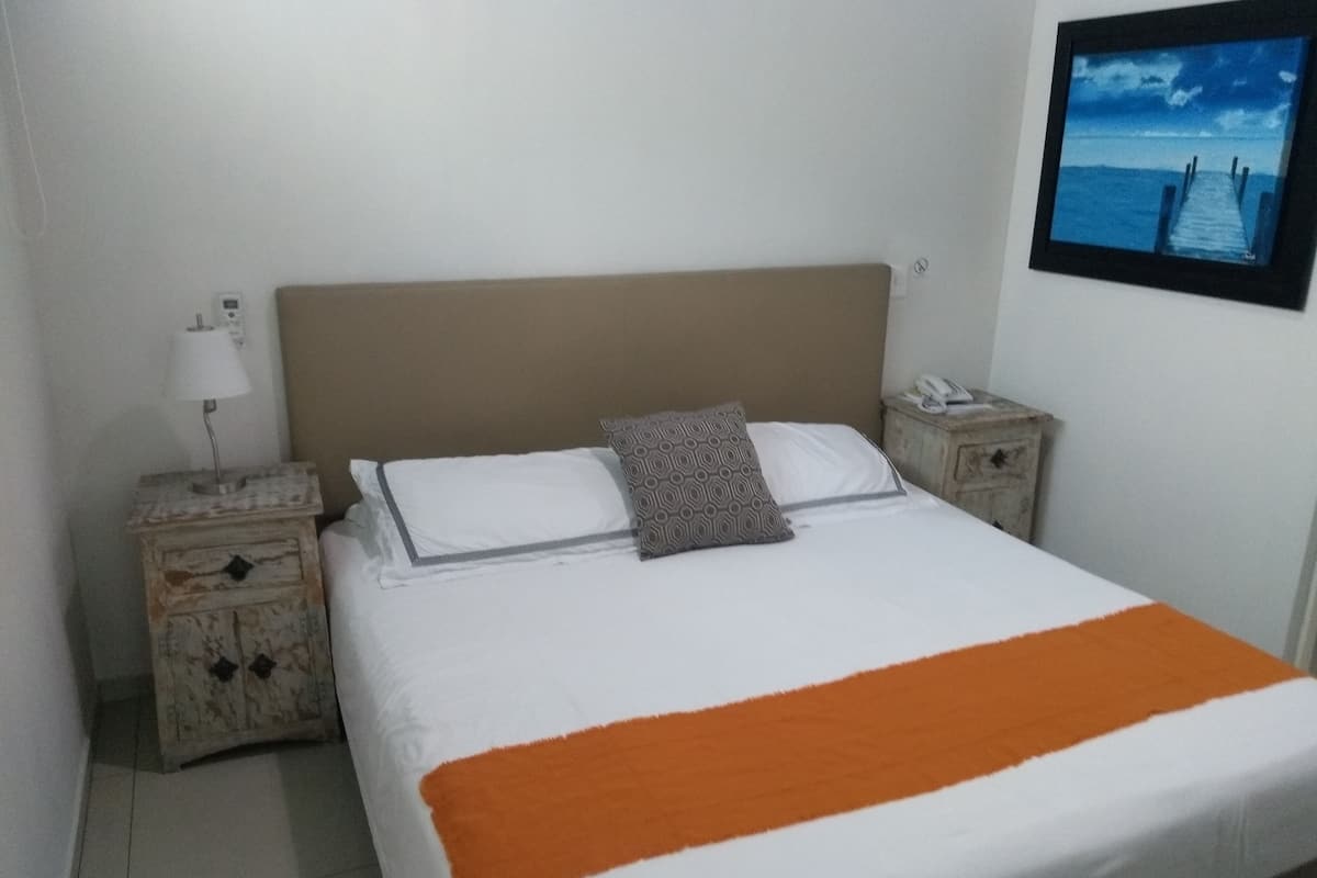 Una cama bien hecha con colcha blanca y naranja, un cojín estampado y una mesita de noche con lámpara y teléfono.