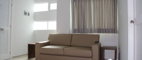 Ein modernes Wohnzimmer mit einem beigefarbenen Sofa, einem kleinen Holztisch und einem großen Fenster mit transparenten Vorhängen.