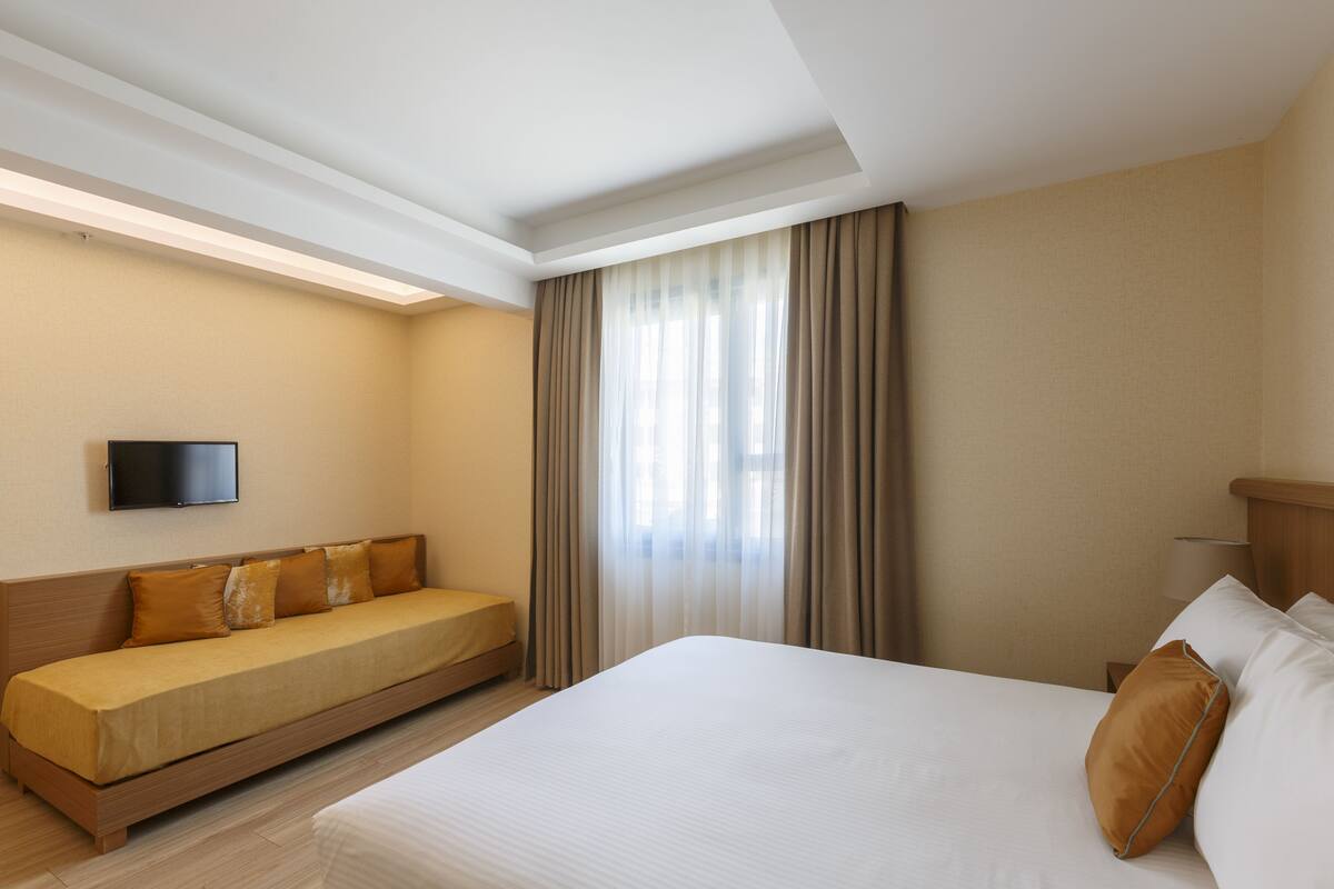 A hotel room with a large bed, a sofa bed, a flat-screen TV mounted on the wall, and a window with curtains.