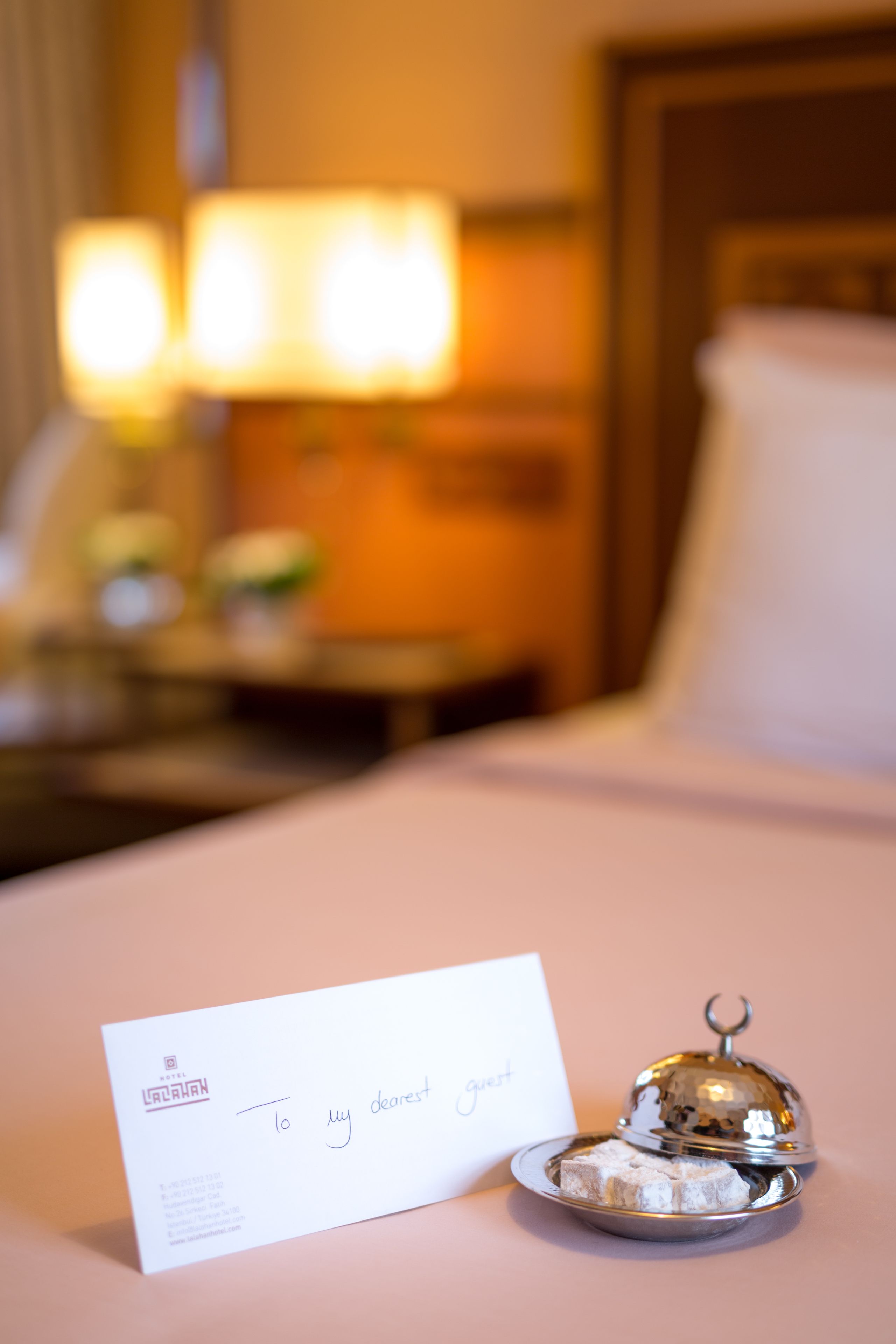 A hotel room with a neatly made bed, a bedside table with a lamp, and a sign reading "To my dearest guest".