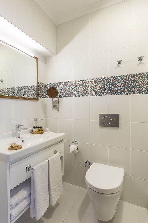 Een badkamer met een wit wastafel, spiegel en toilet. Er is een betegelde rand met patroon boven de wastafel en een handdoekhouder met handdoeken.