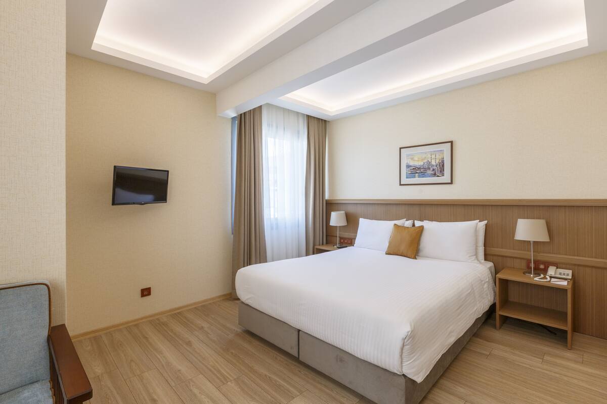 A hotel room with a large bed, a flat-screen TV mounted on the wall, a bedside table with a lamp, and a painting on the wall.