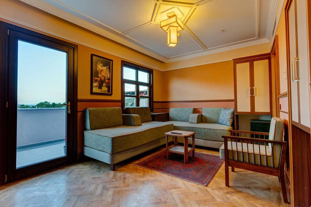 A living room with a sofa, a small table, and a wooden chair. There is a large window and a door leading to a balcony.