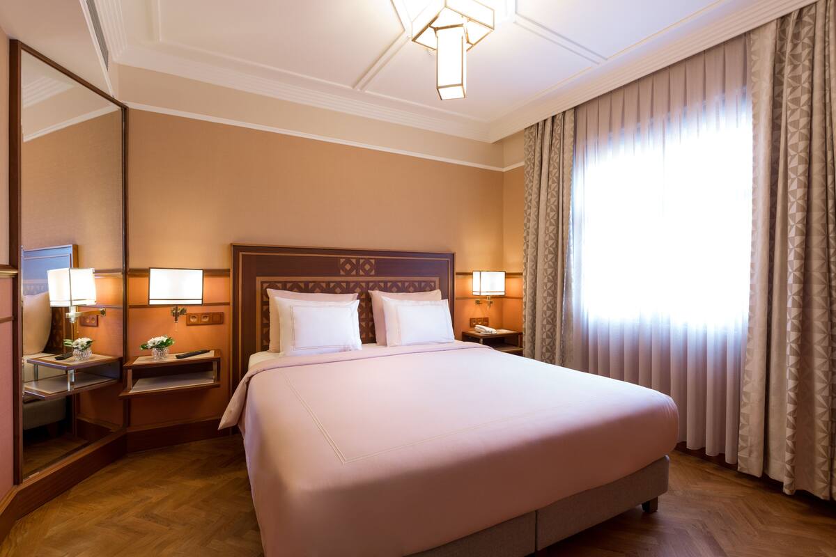 A hotel room with a large bed, bedside tables, lamps, a mirror, and a window with curtains.