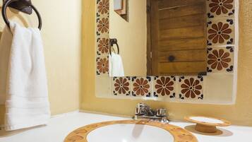 Un baño con tocador de madera, un lavamanos blanco, un espejo con detalle de azulejos florales y una toalla colgada en un aro metálico.