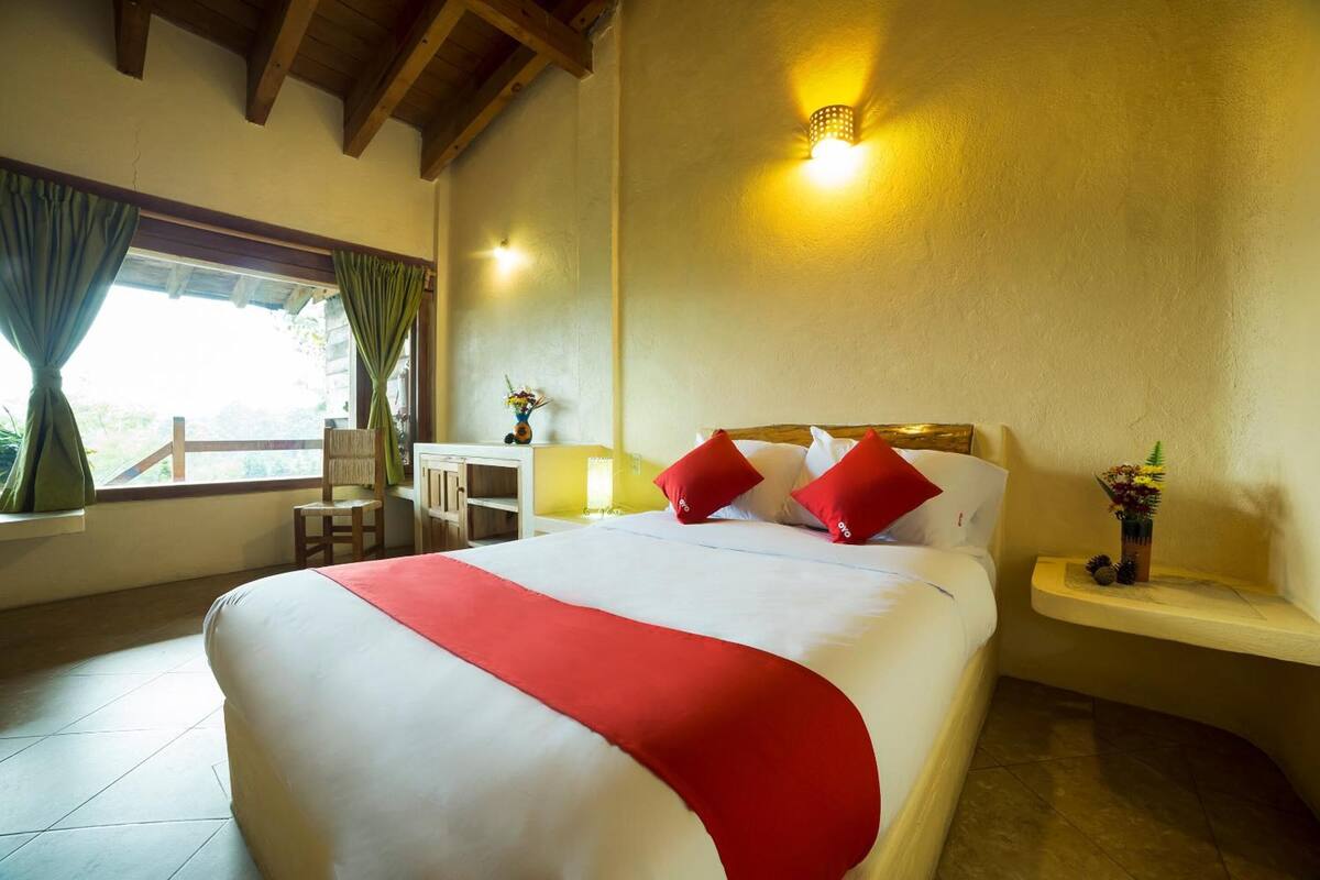 A neatly made bed with a red accent, a bedside table with a vase of flowers, and a window with curtains.