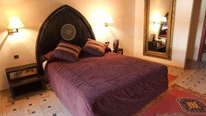 A bedroom with a large bed, two bedside lamps, a nightstand, a mirror, and a patterned rug. - Ouarzazate Le Tichka (Ouarzazate)