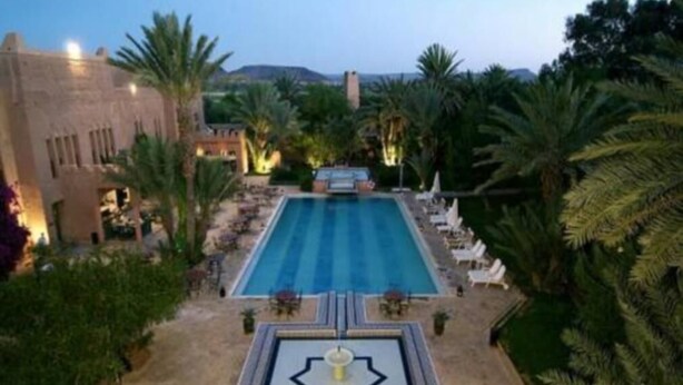 A swimming pool with lounge chairs and a fountain, surrounded by palm trees and a desert landscape. - Ouarzazate Le Tichka (Ouarzazate)
