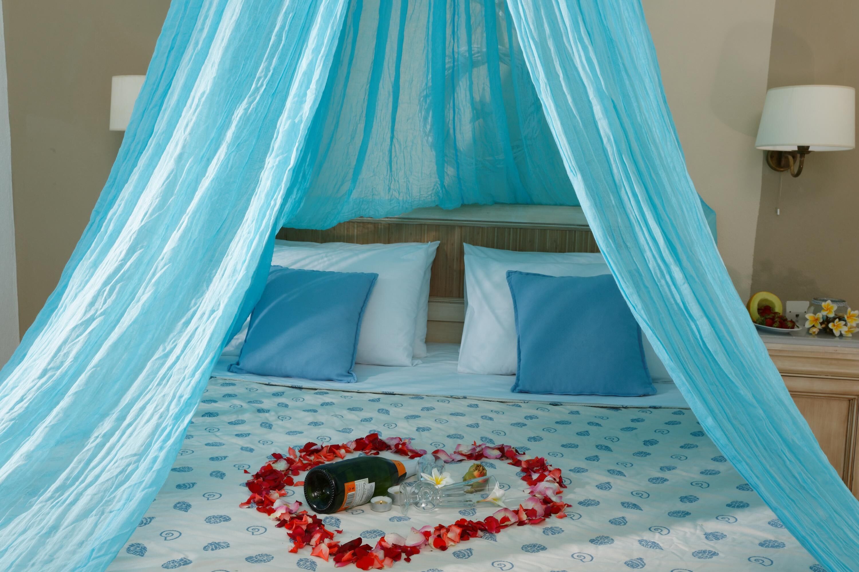 Una cama con dosel, almohadas, una botella y un arreglo de pétalos de rosa con forma de corazón.