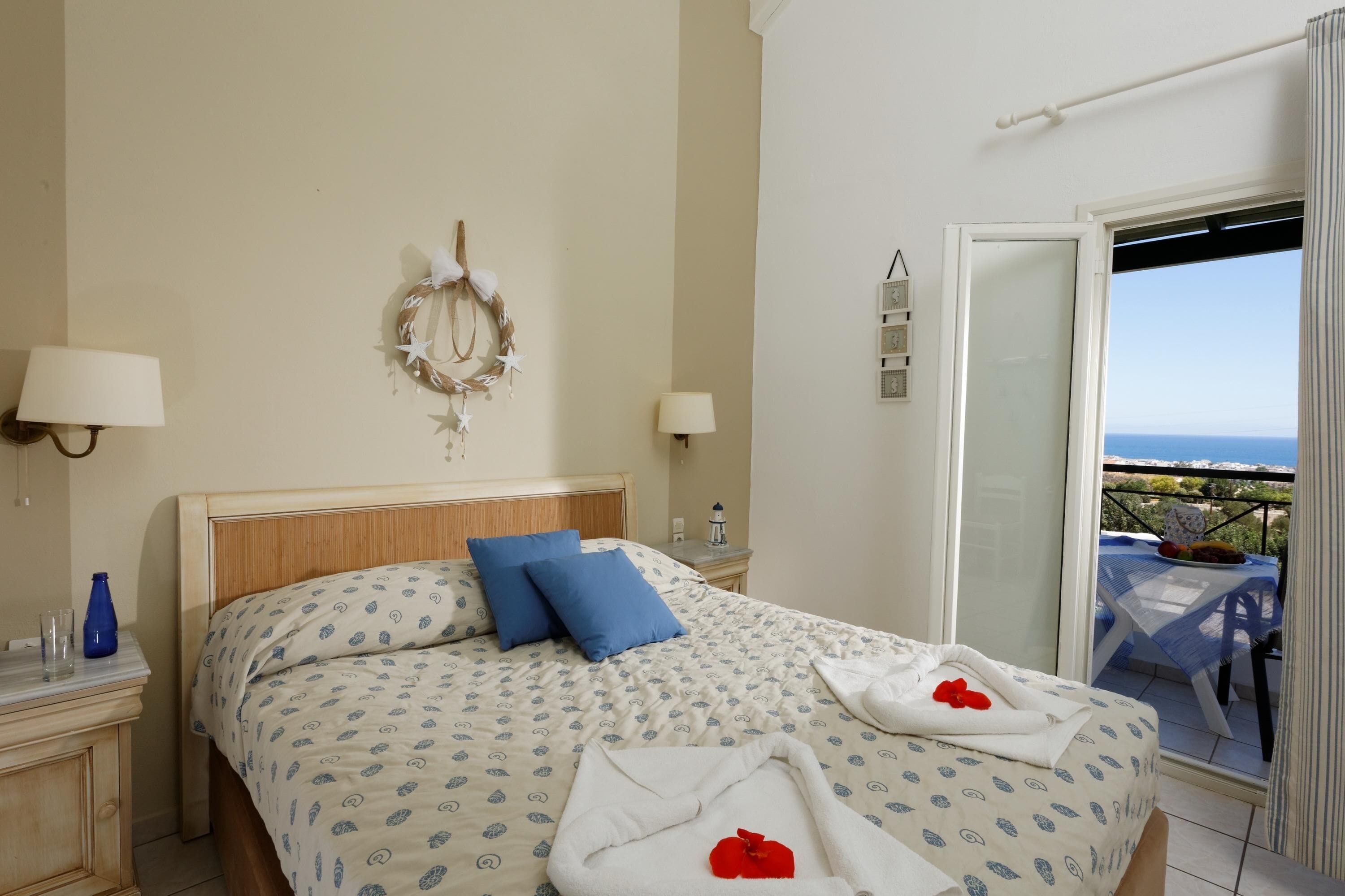 Et hotelværelse med en seng, to sengeborde med lamper, en pyntelig krans, et lille bord med lampe, samt en balkon med bord og stole.