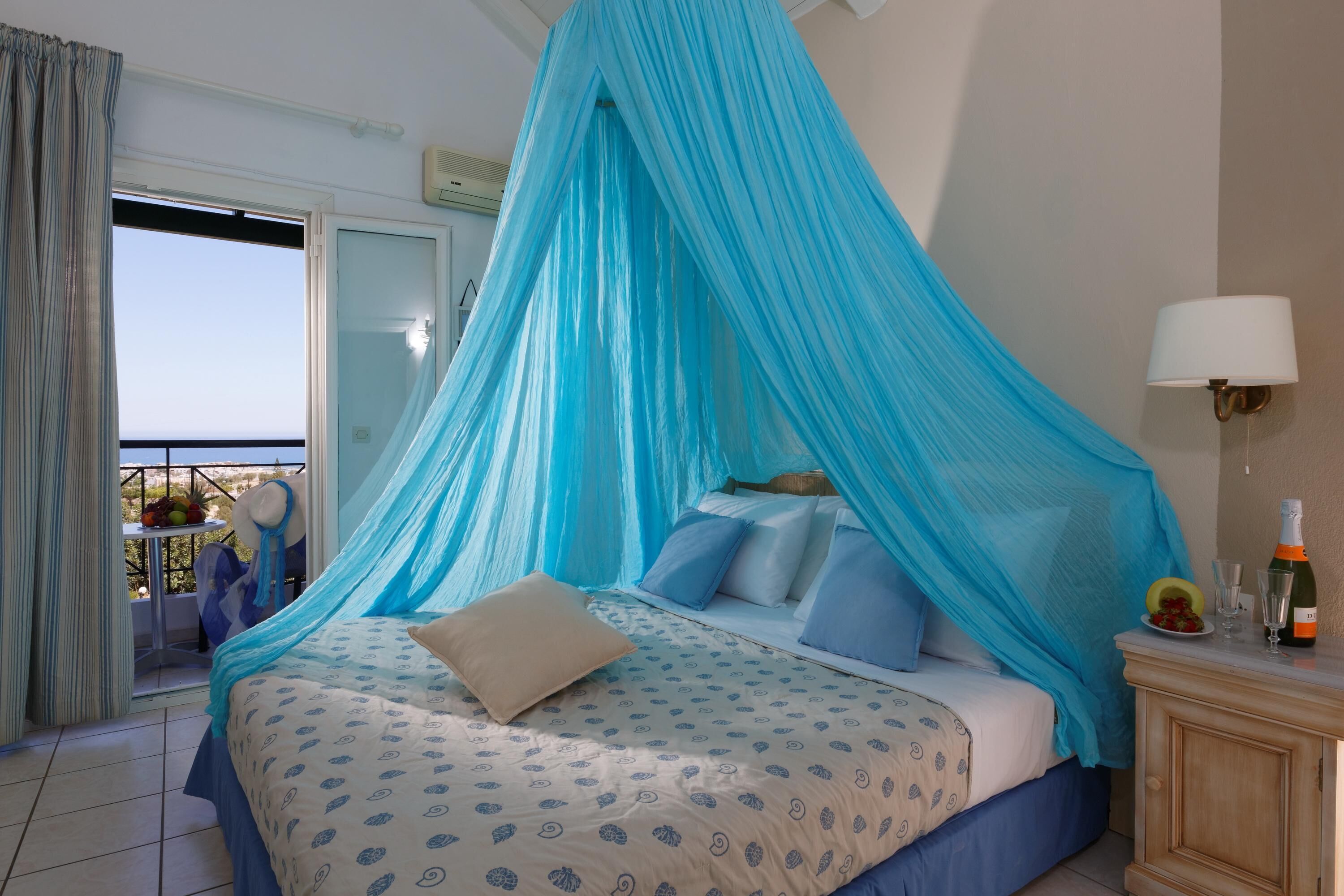 Un dormitorio con una cama bajo un dosel azul, una mesita de noche con una botella y vasos, y vistas al exterior a través de una puerta corrediza.