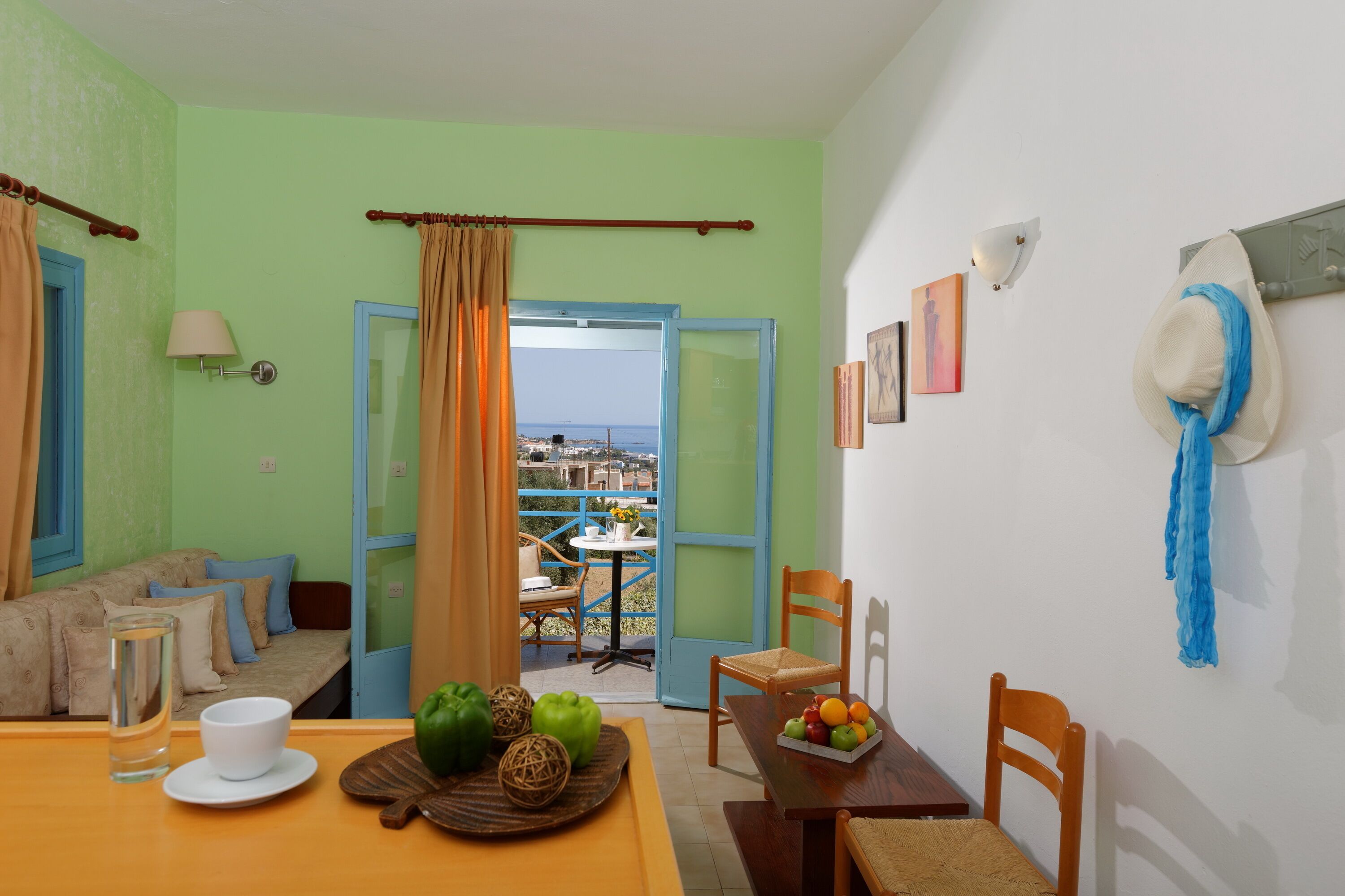 A room with a dining table set with a bowl of fruit, a glass of water, and a plate. There is a chair and a window with curtains.