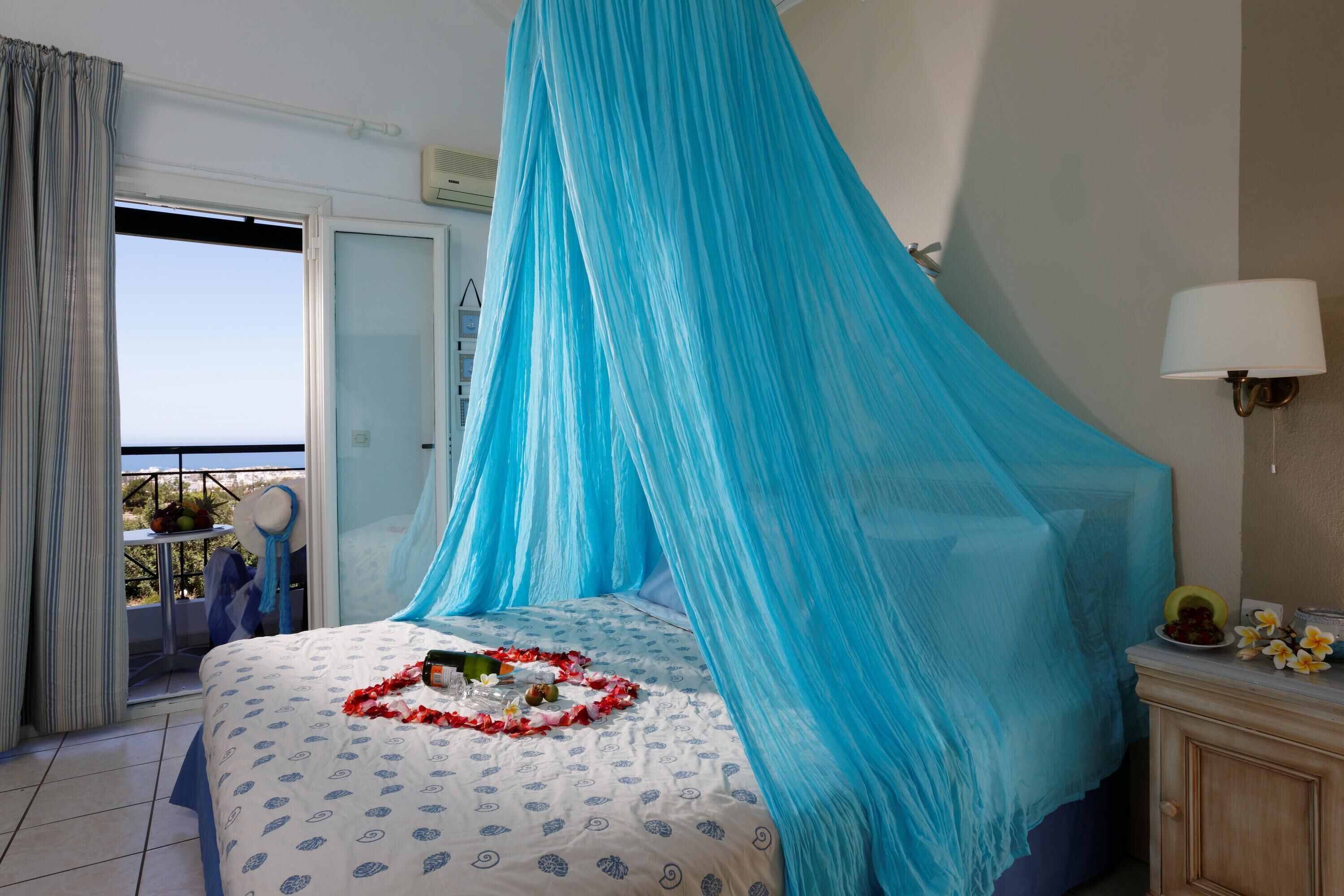 A bedroom with a bed covered by a blue canopy, a bedside table with a lamp, and a view of the outdoors.