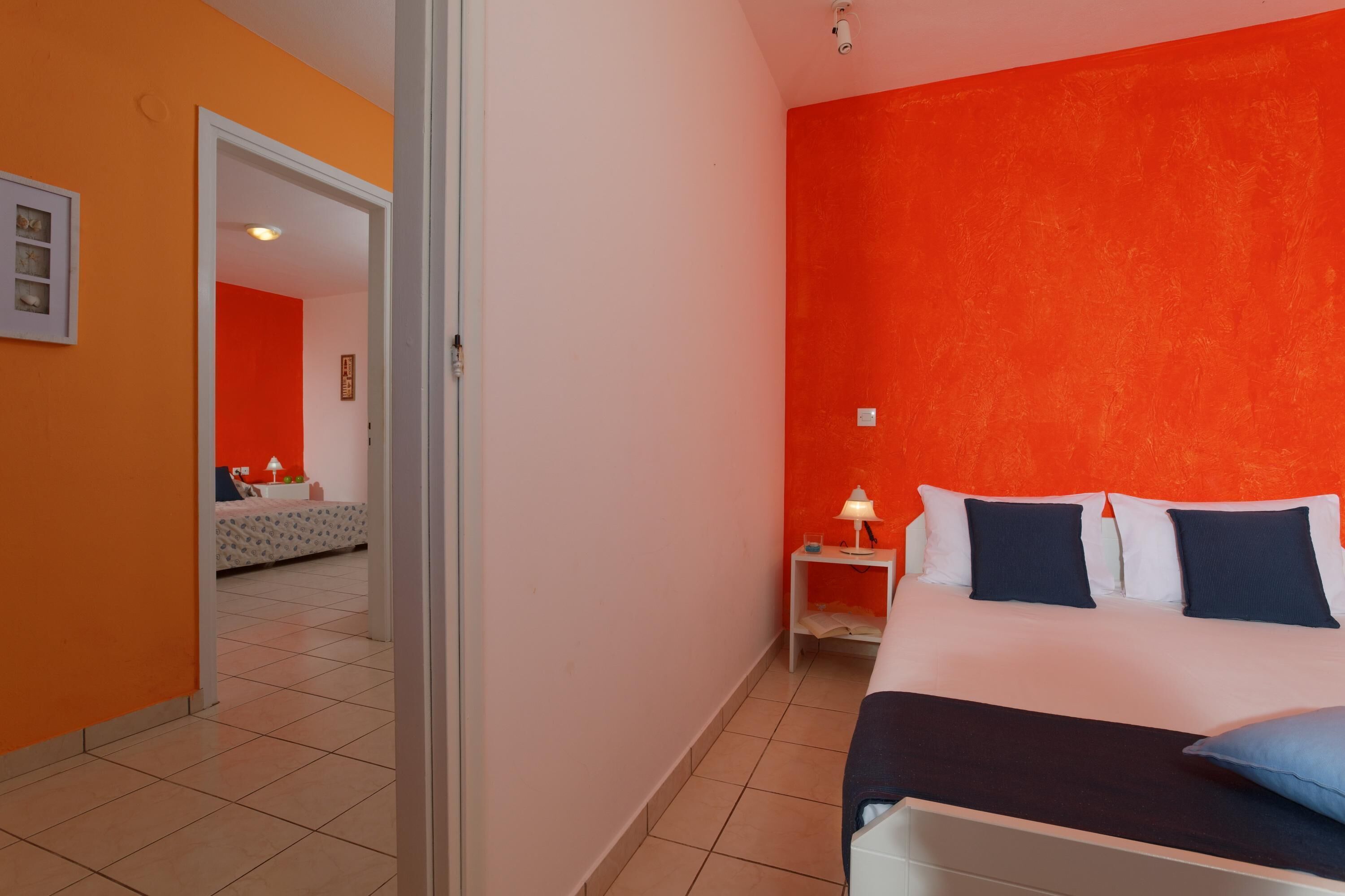 Habitación de hotel con pared de acento anaranjada, cama con ropa de cama blanca y azul oscuro, y mesita de noche con lámpara.