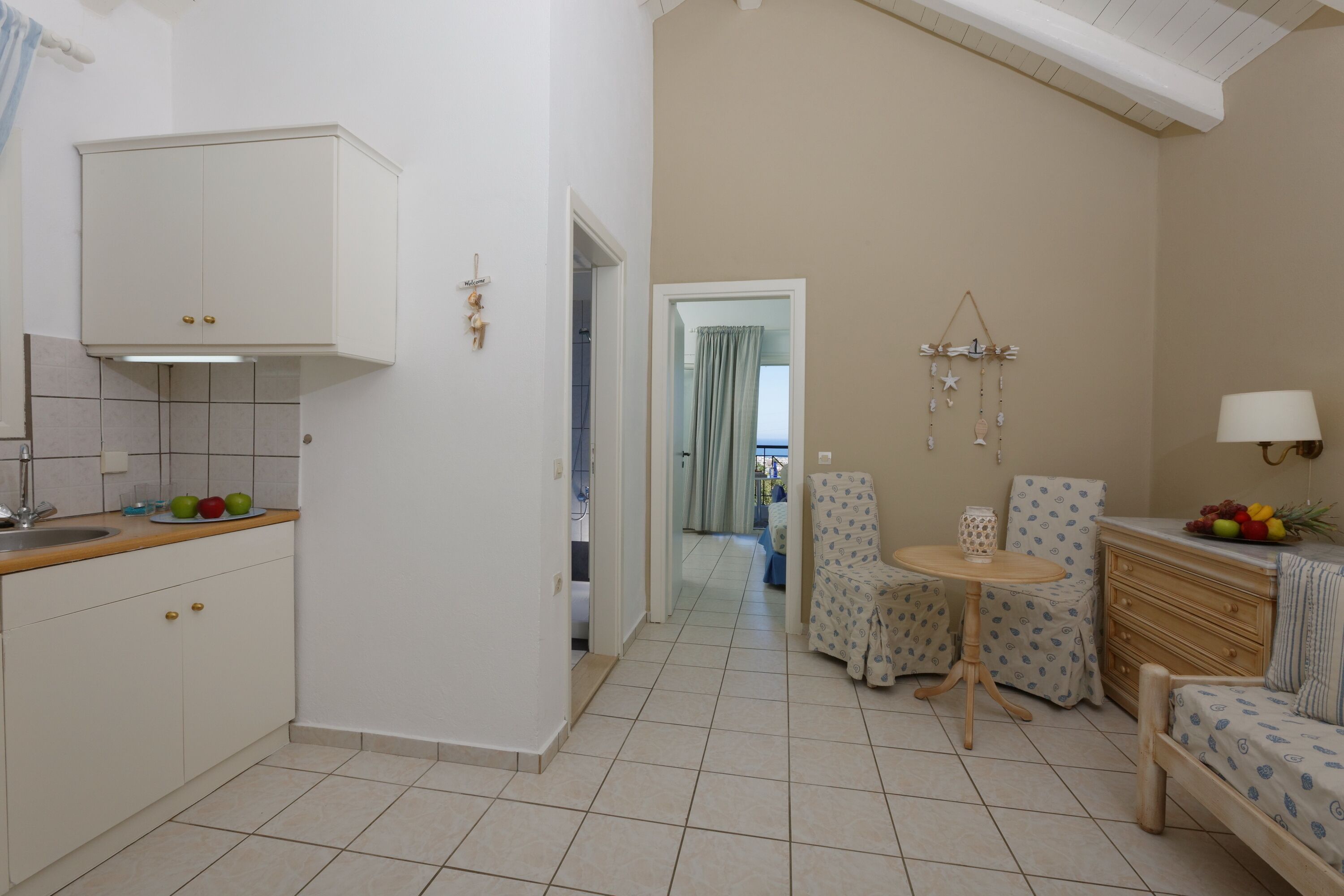 A compact kitchen with a sink and cabinets, a dining area with a table and chairs, and a living space with a sofa and a lamp.