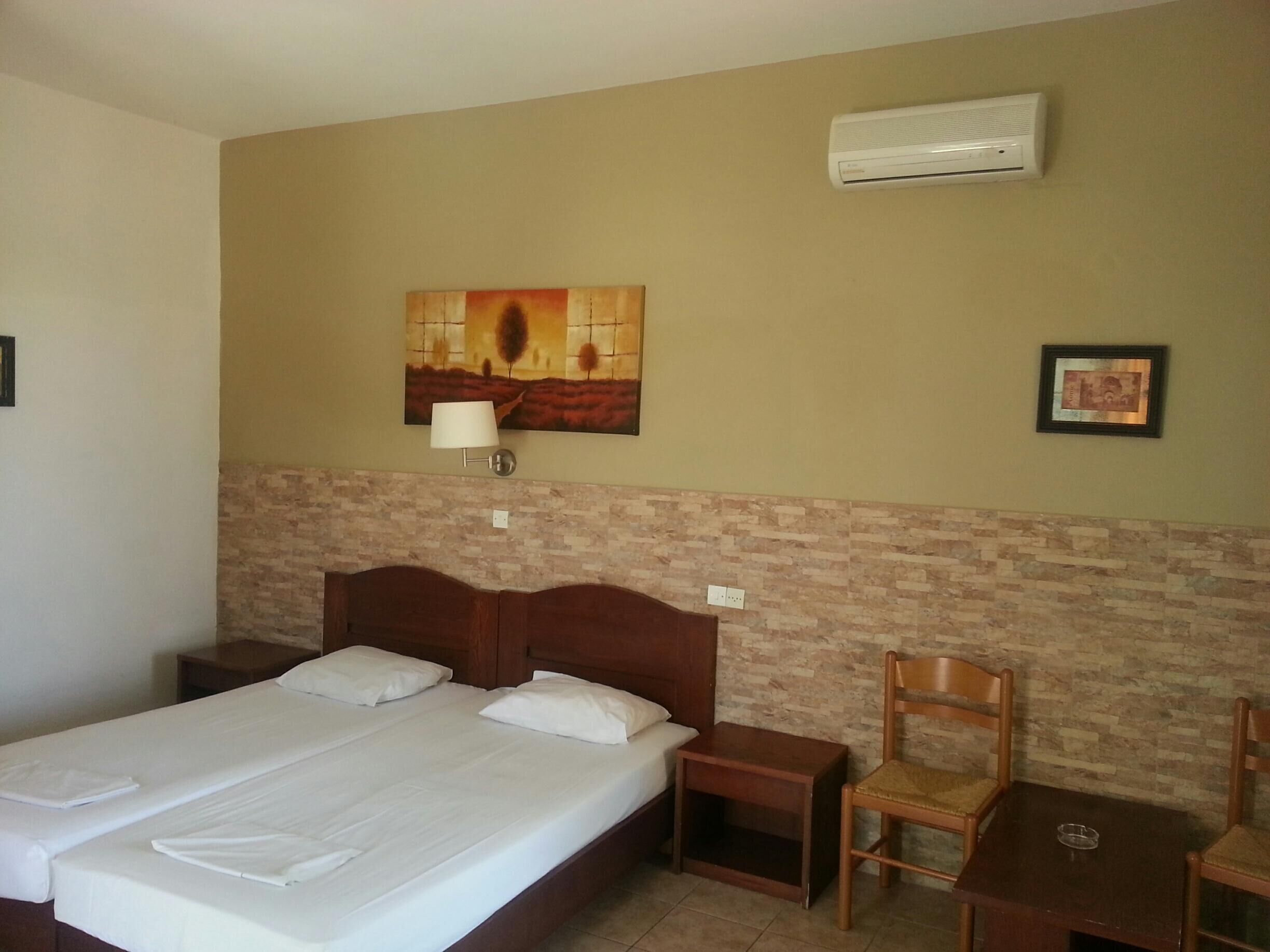 A hotel room with a large bed, a nightstand, a chair, a small table, an air conditioning unit, and a wall-mounted lamp.
