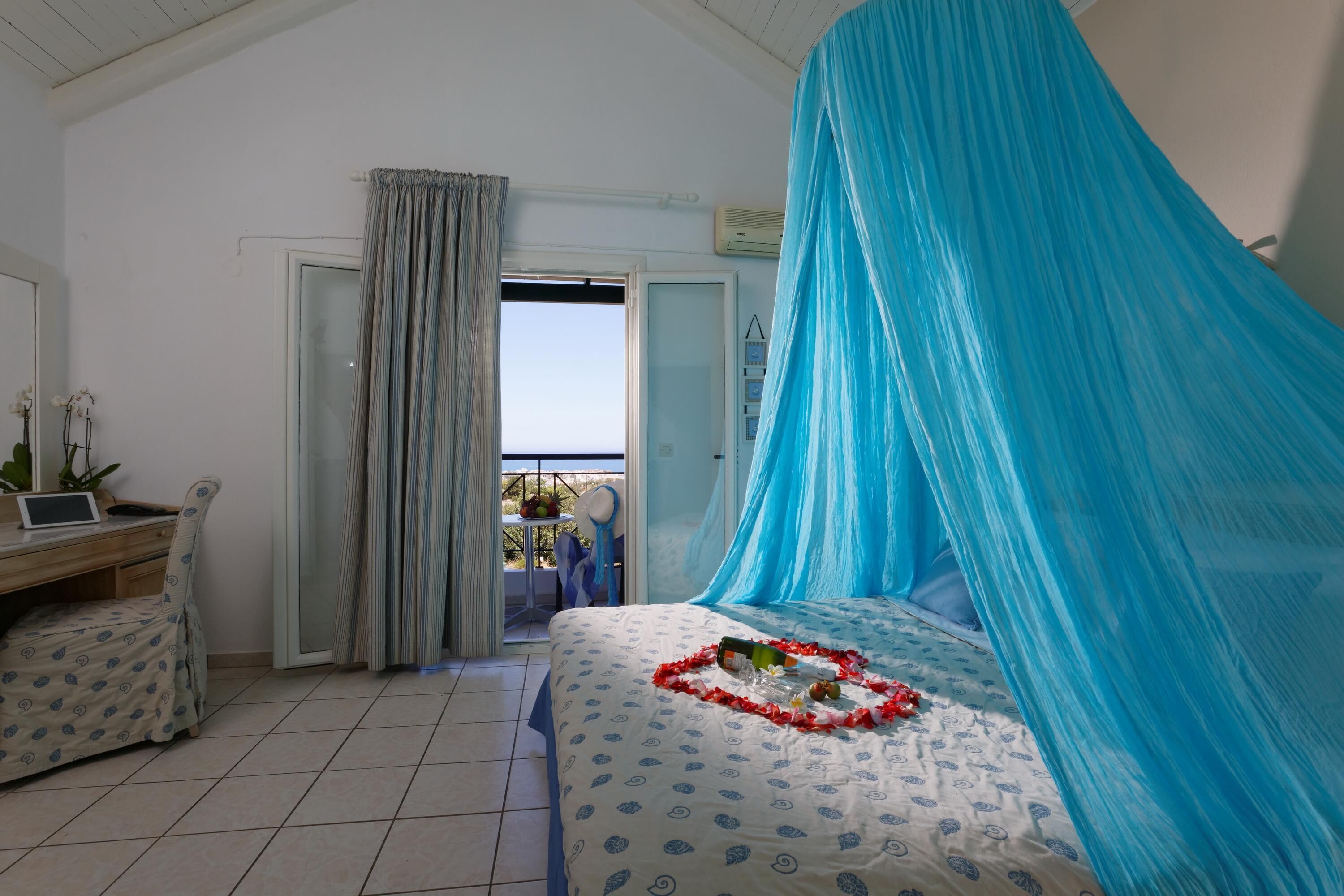 A bedroom with a bed covered by a blue canopy, a desk with a laptop, and a view of the outdoors through a door.