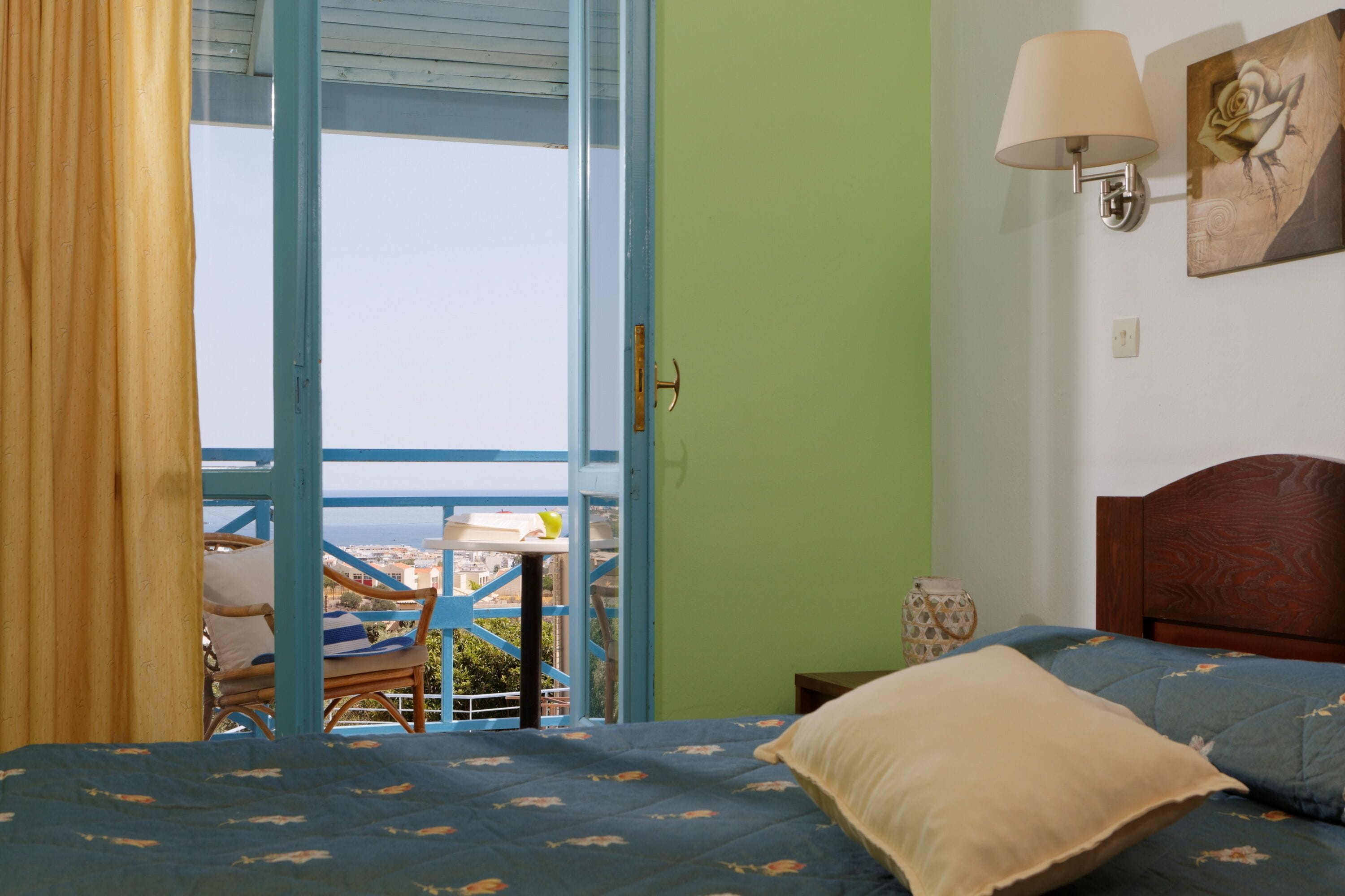 A bedroom with a bed, a nightstand, a lamp, a painting, and a view of the city through an open door.
