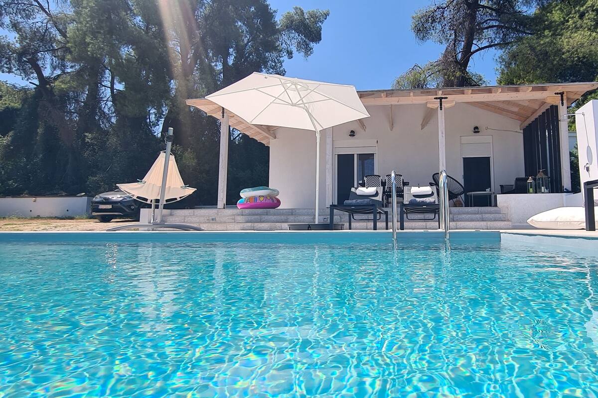 A swimming pool with a covered seating area, surrounded by trees and a white building.