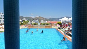 Una piscina con gente, rodeada de sillones de playa y sombrillas, con edificios y una colina al fondo.