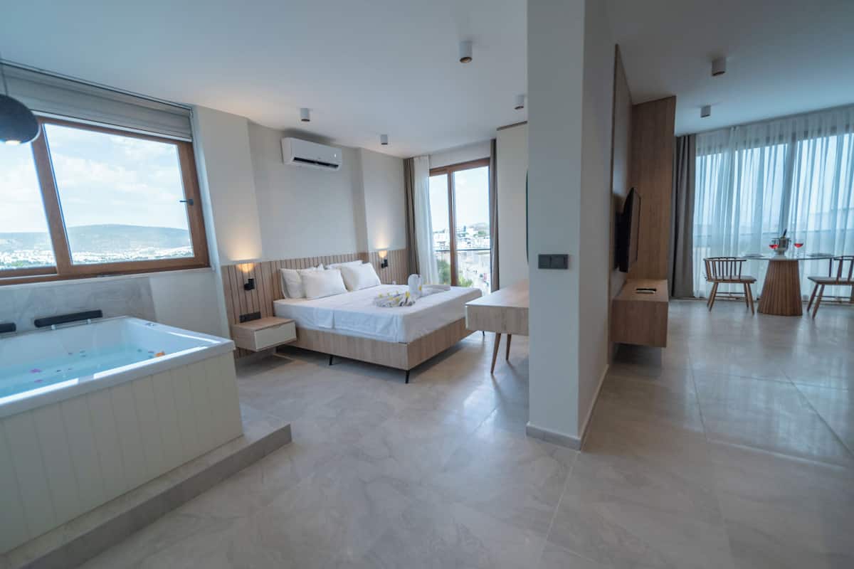 A modern hotel room with a large bed, a sitting area with a table and chairs, a bathtub, and a view of the city.