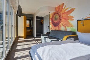 Habitación de hotel moderna con una cama grande, un sofá, una pared con un mural de girasoles y vistas a través de la puerta de vidrio.