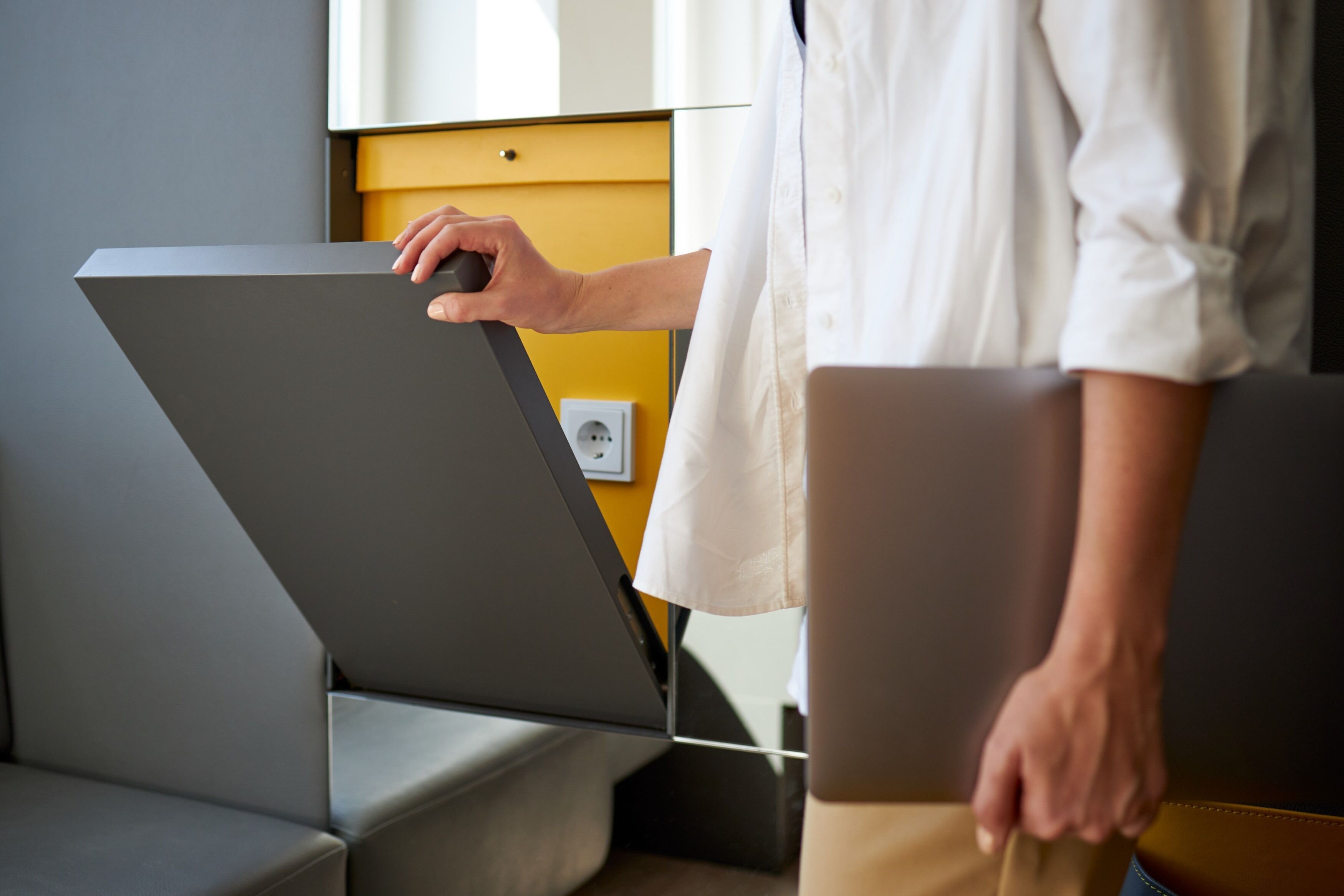 A person in a white shirt interacting with a modern, gray, wall-mounted device, possibly a control panel or interactive display, in a room with a yellow accent wall and a visible electrical outlet.