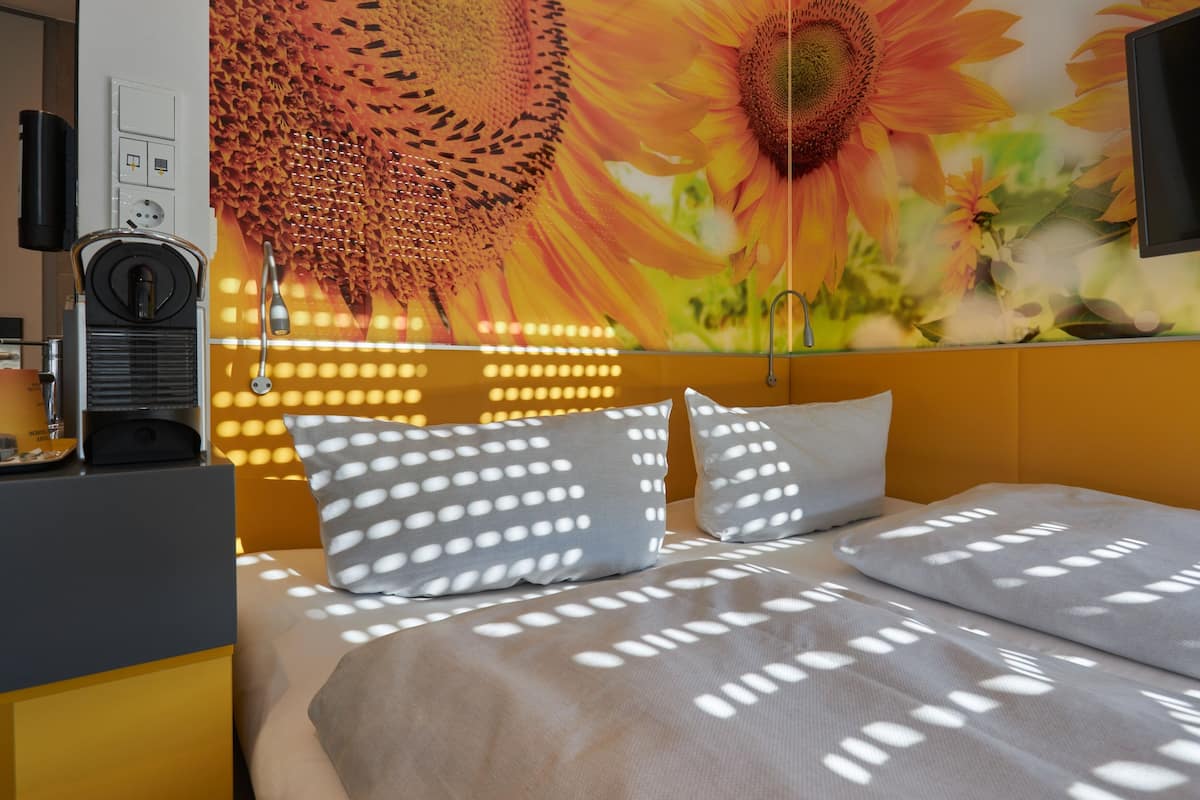 Une chambre d’hôtel avec une grande fresque murale représentant des tournesols, un lit agrémenté d’oreillers à motifs et une machine à café posée sur une table d’appoint.