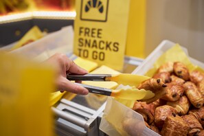 Una mano sosteniendo unas pinzas sobre una bandeja con comida, con un letrero al fondo que dice 'FREE SNACKS TO GO'.