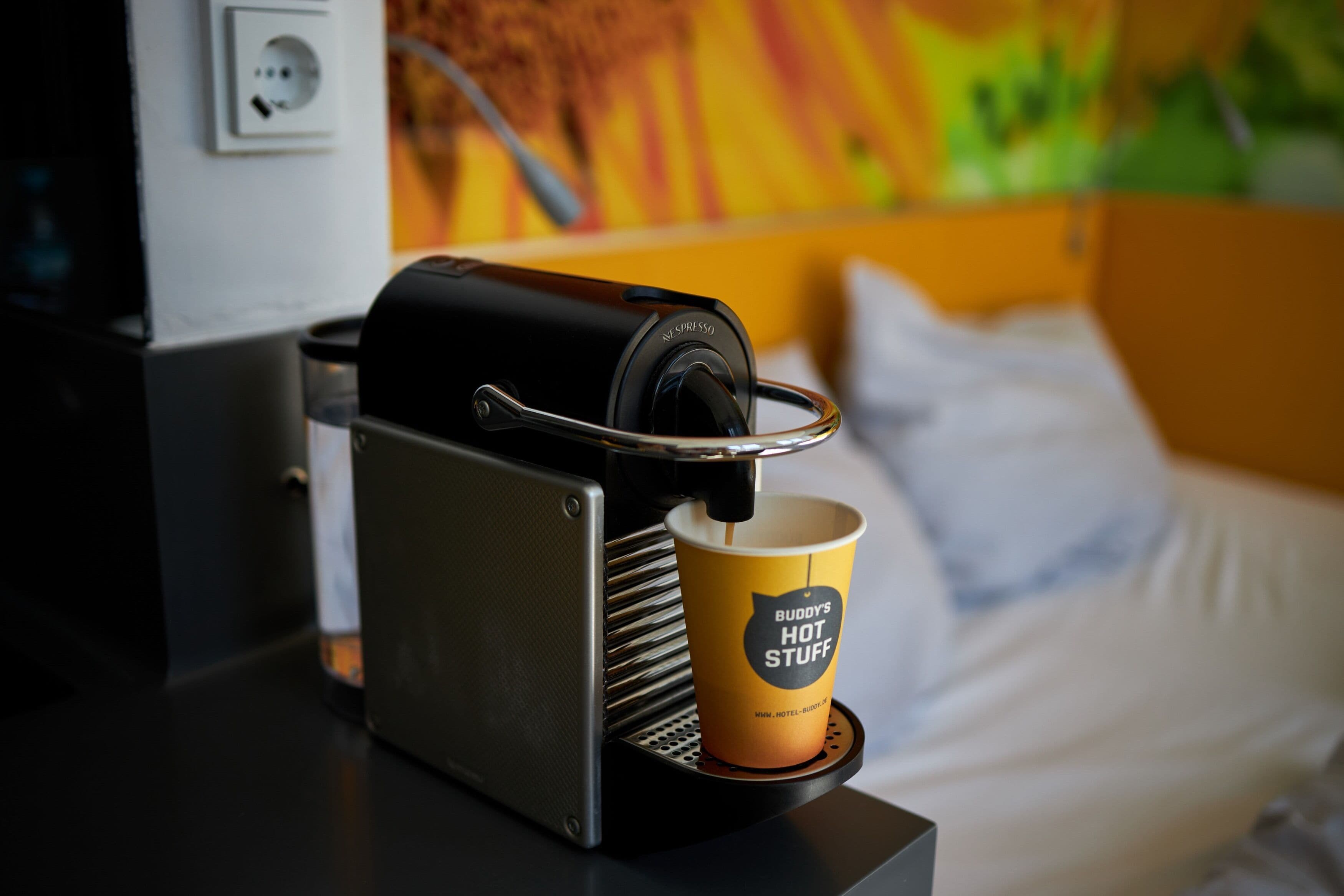 A coffee machine with a cup of coffee on a tray, a power outlet, and a bed with pillows in the background.