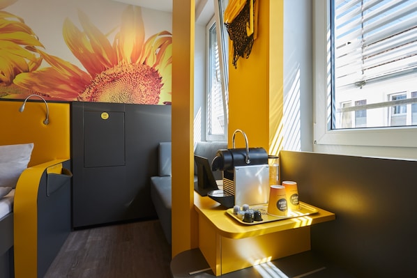 Una habitación pequeña con cama, una mesita con cafetera y ventana con persianas.