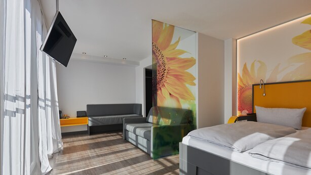 A modern hotel room with a large bed, a sofa, a TV mounted on the wall, and a large window with curtains. - Buddy Hotel (Munich)