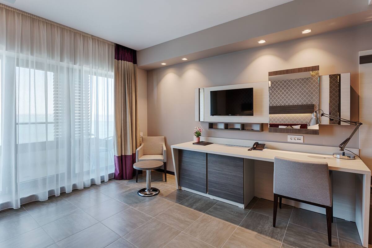 A modern hotel room with a large window, a desk with a chair, a TV, and a mirror.