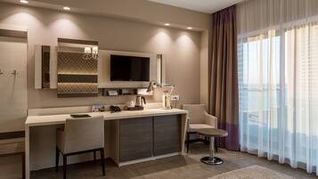 A modern hotel room with a desk, two chairs, a TV, and a large window with curtains.