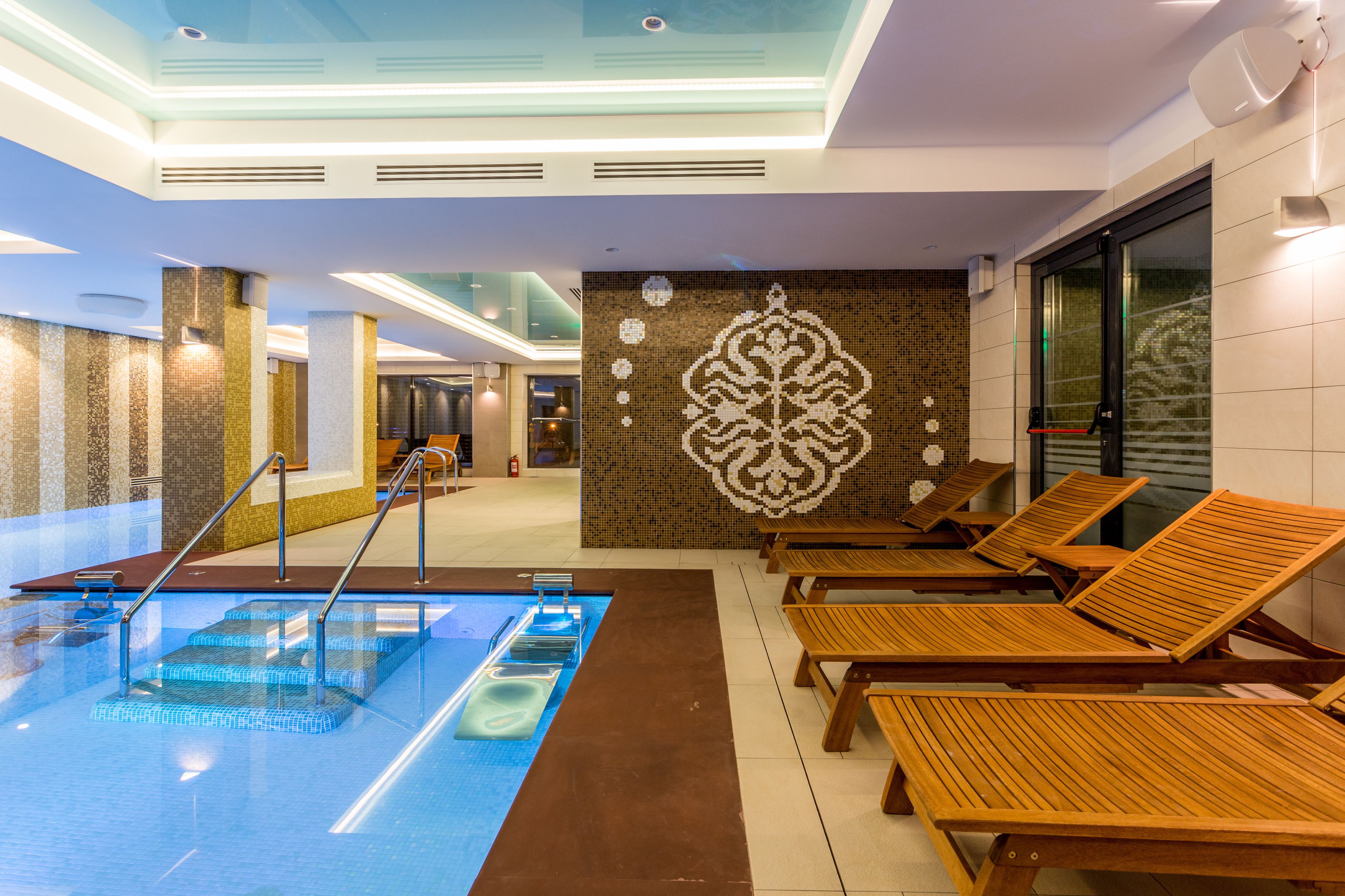 A modern indoor pool area with a glass ceiling, a mosaic wall, and wooden lounge chairs.