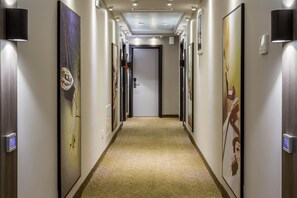 Corredor de hotel com obras de arte nas paredes, piso coberto por carpete e uma porta no final.