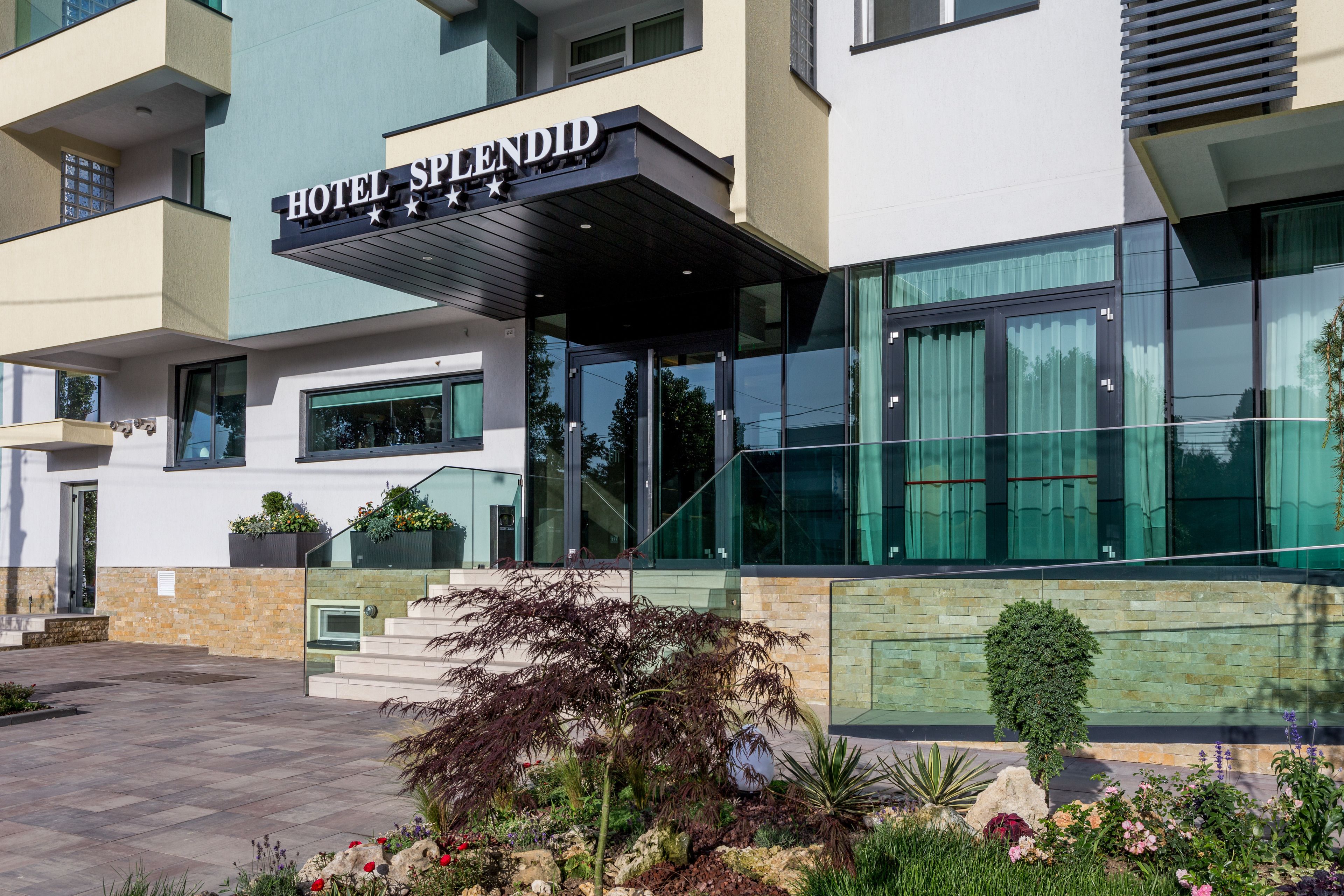 A modern hotel entrance with a glass facade, a sign reading "HOTEL SPLEENDID," and a landscaped garden area with various plants and flowers.