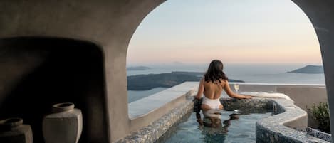 En kvinna i en vit baddräkt sitter i en infinitypool med stenväggar, och från poolen har man utsikt över havet och det avlägsna landskapet.
