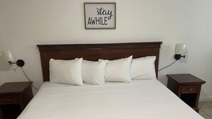 A neatly made bed with white linens and a floral throw blanket, wooden headboard, bedside tables with lamps, and a 'stay AWhile' sign on the wall.