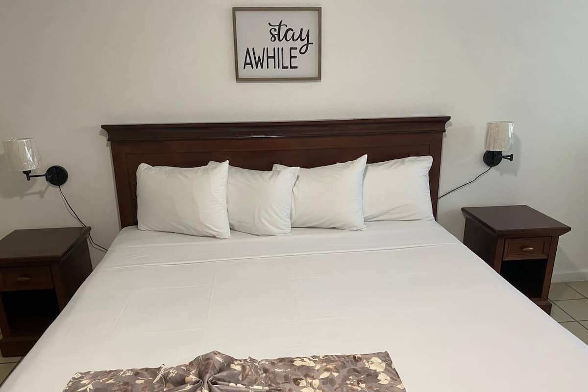 A neatly made bed with white linens and a floral throw blanket, wooden headboard, bedside tables with lamps, and a 'stay AWhile' sign on the wall.