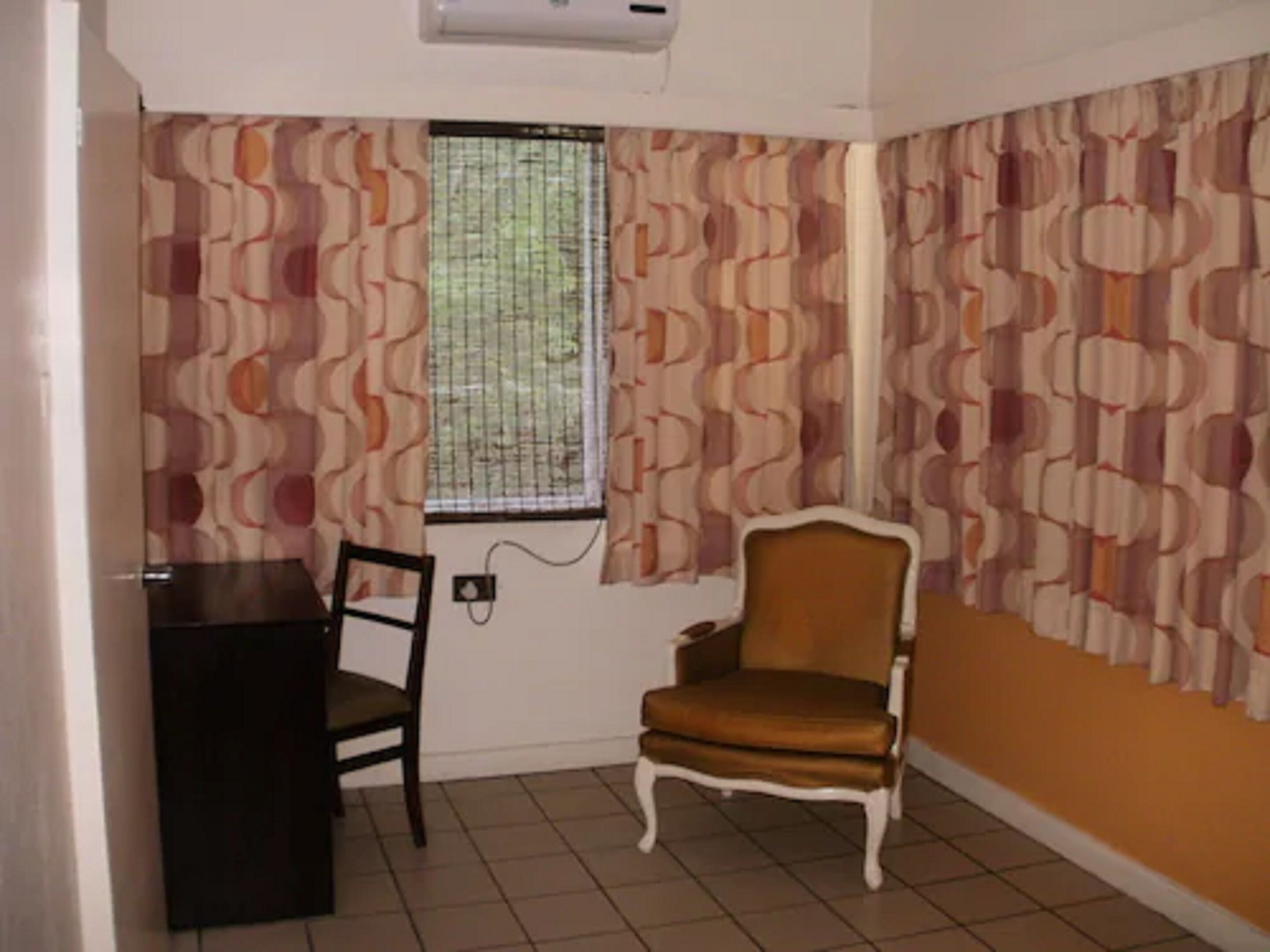 Une pièce avec un papier peint à motifs, une fenêtre avec des barreaux en métal, un fauteuil marron et un bureau noir.