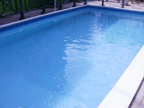 A rectangular swimming pool with clear blue water, surrounded by a tiled deck and a green metal fence.