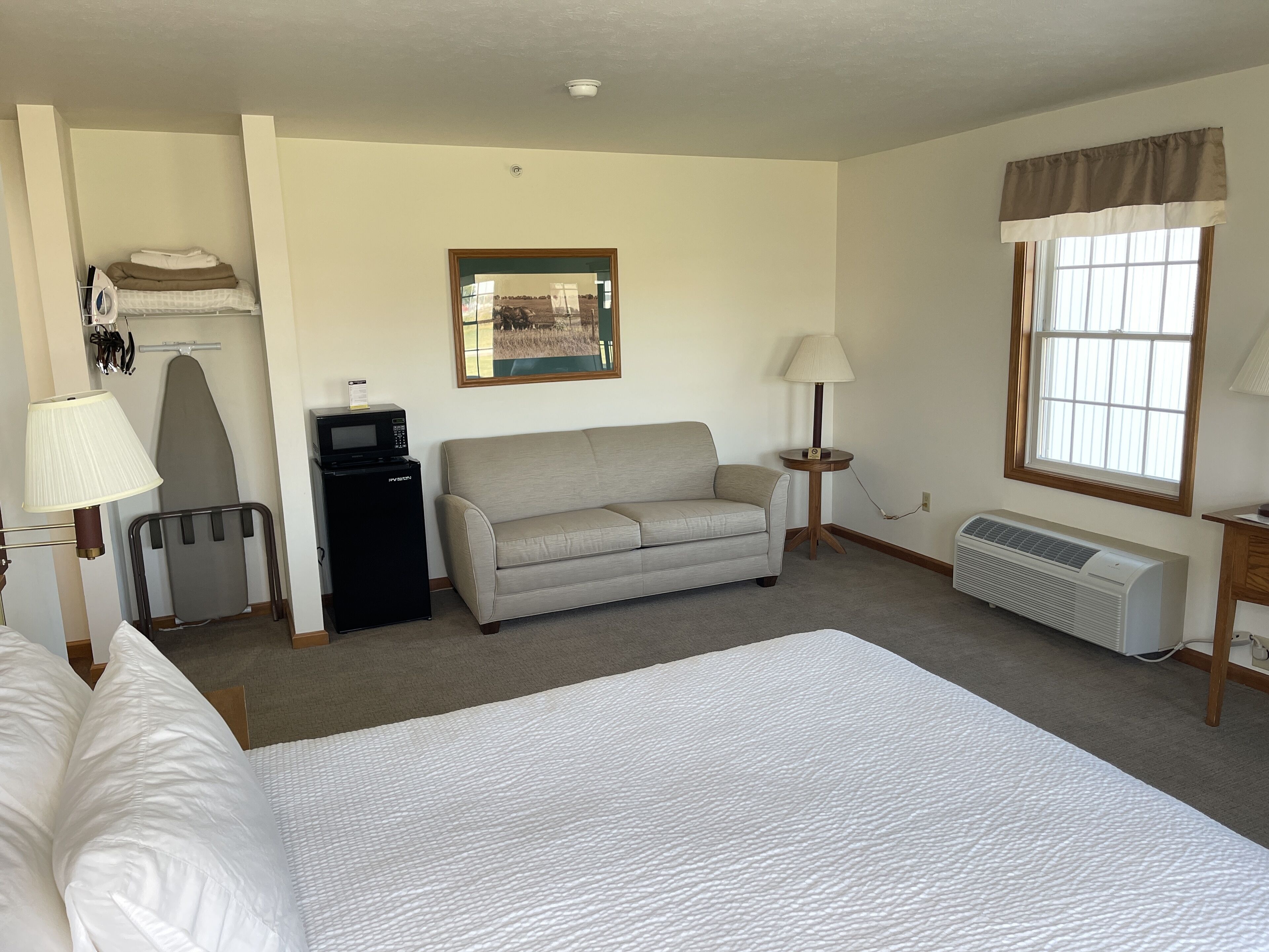 A hotel room with a bed, a sofa, a desk with a computer, a lamp, an ironing board, and a window with curtains.