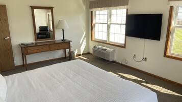 A bedroom with a large bed, a wooden desk with a lamp, a mirror, a wall-mounted TV, and a window with curtains.