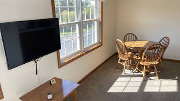 A room with a TV mounted on the wall, a small table with a phone, a dining table with chairs, and a window with a view of trees and a parking lot.
