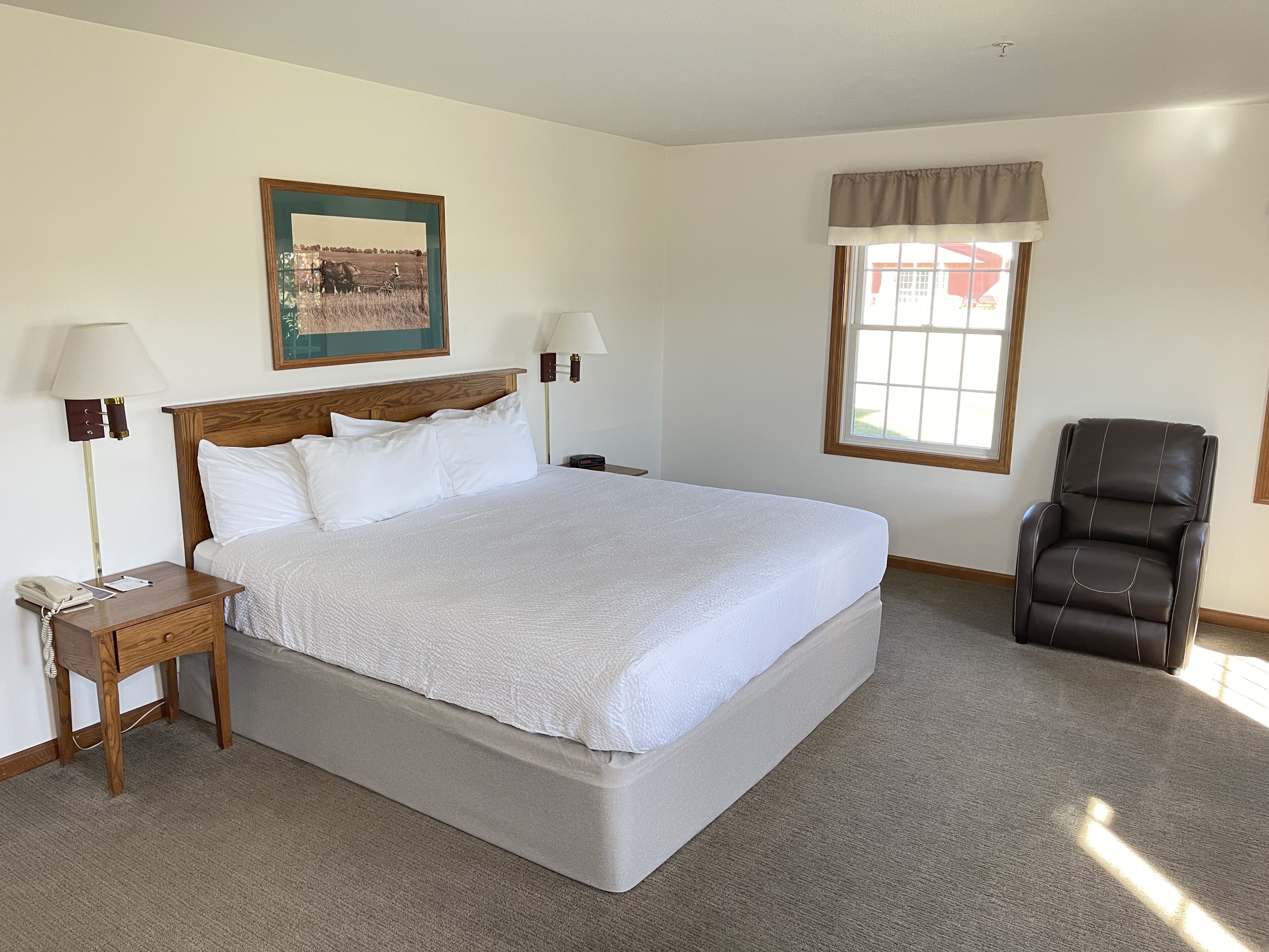 A neatly arranged bedroom with a large bed, bedside lamps, a chair, a window with curtains, and a framed picture on the wall.