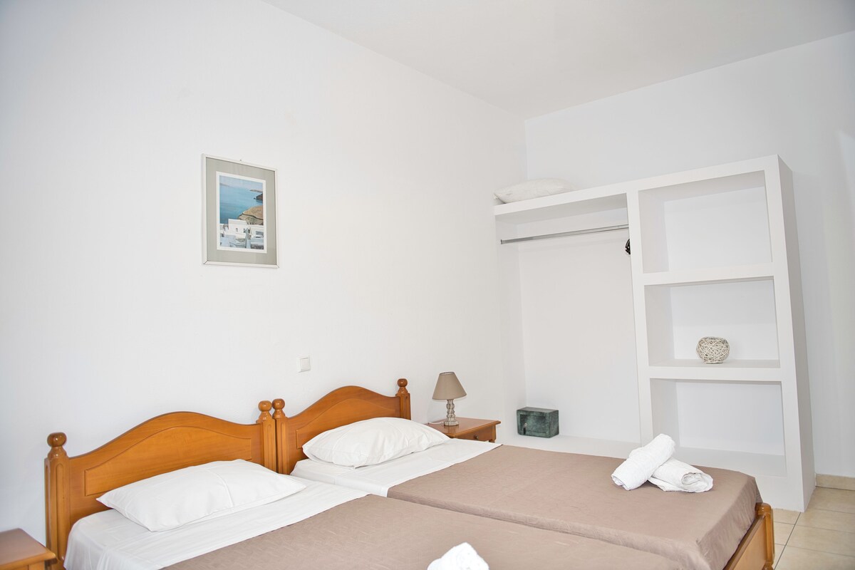 A double bed with a wooden headboard, two white pillows, and a beige bedspread. There is a small bedside table with a lamp and a framed picture on the wall.