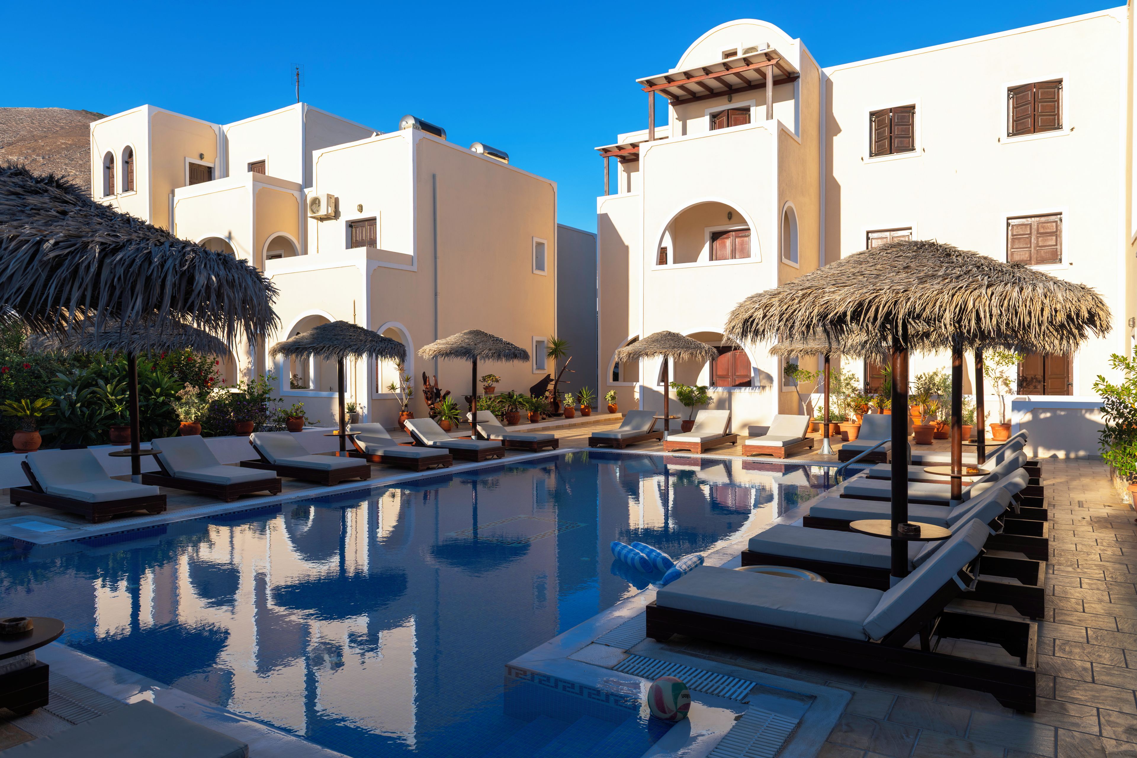 A pool area with lounge chairs and umbrellas, surrounded by buildings with white walls and arched windows.
