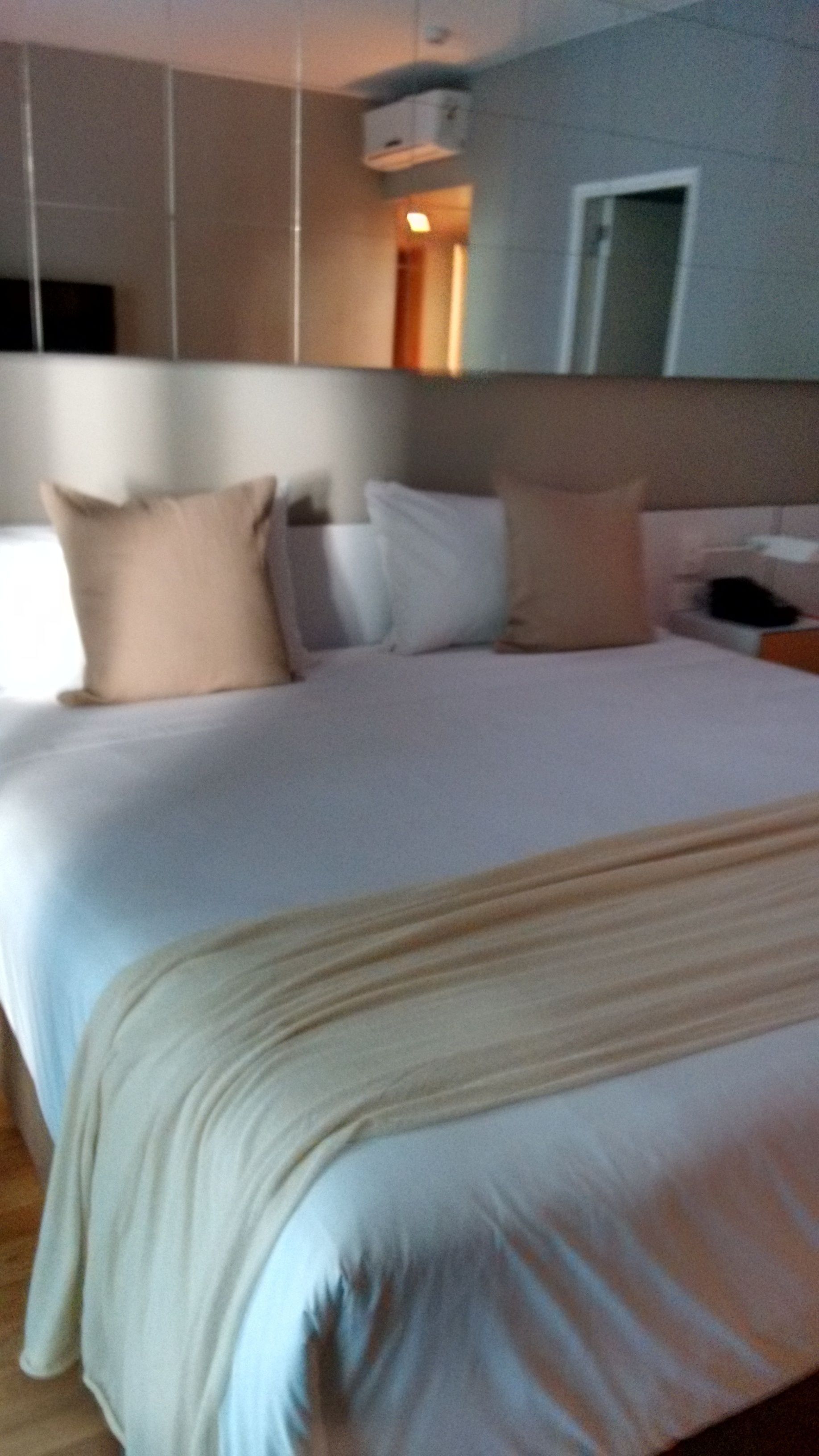 A hotel room with a large bed, multiple pillows, a bedside table, and a wall-mounted air conditioning unit.